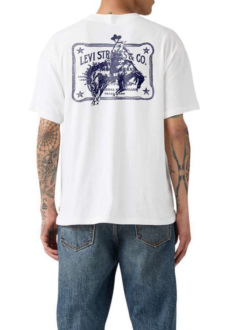 Hombre que lleva una camiseta blanca de Levi's con gráfico de un vaquero montando un caballo en la espalda, combinada con vaqueros azules, mostrando tatuajes en ambos brazos.