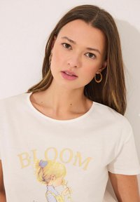 Witte katoenen T-shirt met een afbeelding van een meisje dat een bloem vasthoudt, met het woord "BLOOM" in zachtgeel boven haar gedrukt.