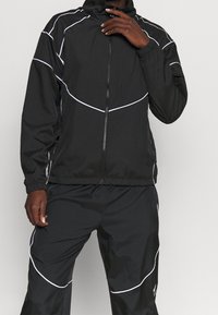 Chaqueta de chándal negra con ribetes blancos, cierre con cremallera y puños elásticos, combinada con pantalones a juego. La tela parece ligera y suave.