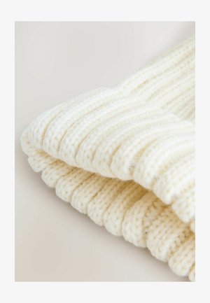 Krämfärgad stickad mössa med en tjock ribbad design. Tillverkad av mjukt garn, har tyget ett mysigt och strukturerat utseende.