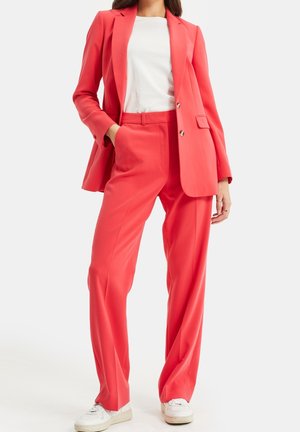 Woman wearing coral pink tailored blazer and matching pants with white sneakers and white crew neck top, hand in pocket, standing on white background.