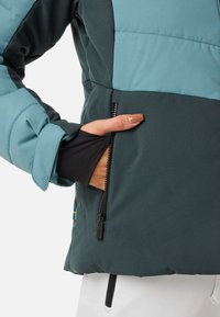 Teddy-turquoise und dunkelgrüne gepolsterte Jacke mit einer seitlichen Reißverschlusstasche, kontrastierenden Stoffen und einem kleinen schwedischen Flaggenakzent in der Nähe des Saums.