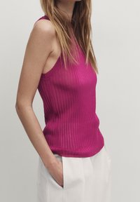 Kobieta w bezrękawnikowej, prążkowanej bluzce w kolorze magenty i białych spodniach, z jedną ręką w kieszeni, na jednolitym jasnym tle.