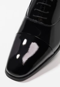 Chaussure Oxford en cuir verni noir avec lacets fermés et une finition brillante qui reflète la lumière sur un fond blanc.