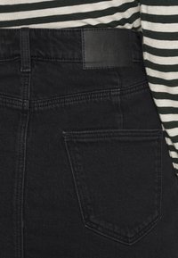 Jeans in denim nero con vita alta, dotati di una toppa in pelle sul retro, tasche standard e cuciture finissime sui bordi. Visibile una maglietta a righe.