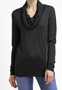 Schwarzes Langarmoberteil mit Schalkragen, aus einem schimmernden Stoff gefertigt. Verfügt über einen seitlichen Reißverschluss und einen lockeren Schnitt, kombiniert mit blauen Jeans.