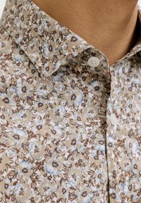 Gros plan sur le col d'une chemise beige à motif floral avec des fleurs bleu clair et marron, présentant des boutons blancs et une zone de peau visible au niveau du cou.