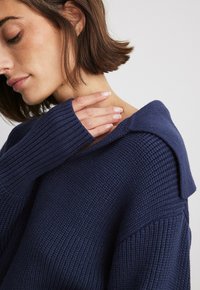 Navy blue knitted sweater with a textured fabric, featuring a folded collar and long ribbed sleeves. Close-up view of neckline and hand.