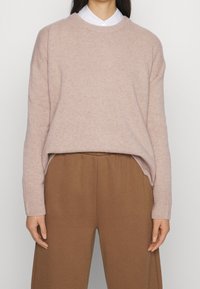 Jasnoróżowy sweter wykonany z miękkiego materiału, okrągły dekolt, opadłe ramiona i ściągacze przy mankietach, połączony z brązowymi, luźnymi spodniami.