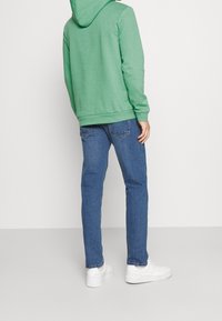 Persona vistiendo una sudadera verde, jeans azules y zapatillas blancas de pie de espaldas contra un fondo claro liso.