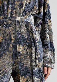 Tela texturada con un patrón floral en tonos azul marino y crema, con una cintura ceñida y mangas anchas, creando una silueta drapeada.