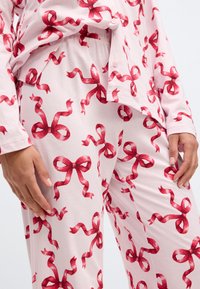 Conjunto de pijama rosa claro com um padrão de fita vermelha repetido; tecido suave, mangas longas, camisa de botão e calças com cintura elástica.