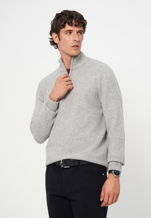 Homme aux cheveux bouclés portant un pull gris clair à fermeture éclair quart de zip et un pantalon noir, ajustant le col du pull avec sa main droite.
