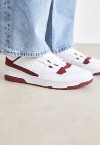 Sneakers bianchi con accenti e logo color marsala, caratterizzati da un upper in pelle testurizzata, punta rotonda e suola in gomma. Cuffs dei pantaloni in denim parzialmente visibili.