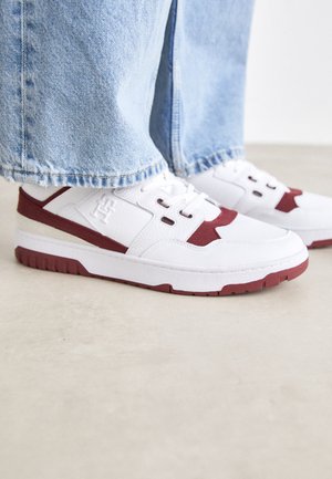 Sneakers bianchi con accenti e logo color marsala, caratterizzati da un upper in pelle testurizzata, punta rotonda e suola in gomma. Cuffs dei pantaloni in denim parzialmente visibili.