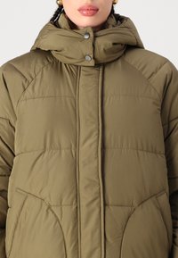 Casaco puffer verde-oliva com gola alta, fecho de botão de pressão e dois bolsos laterais. Apresenta textura acolchoada e detalhes costurados.