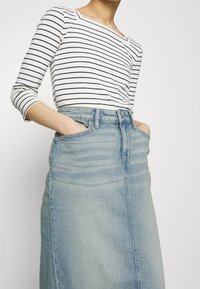 Bílá blůza s dlouhými rukávy a námořnickými vodorovnými pruhy, spárovaná se sukní z lehkého modrého denimu s mírným vzestupem, která má boční kapsy a rovný střih.