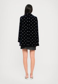 Frau mit langen, dunklen Haaren, die eine schwarze, floral gemusterte Jacke, einen kurzen grauen Rock und weiße, hochhackige Mules trägt, blickt von einem einfarbigen Hintergrund weg.