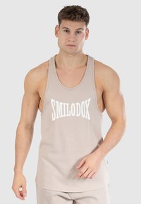 Beige tanktop met witte "SMILODOK" logo, gemaakt van zachte stof, met brede armsgaten en een relaxte pasvorm, ideaal voor actieve kleding.