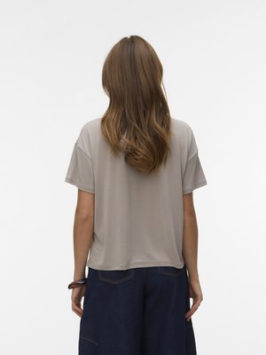 Γυναίκα με μακριά κυματιστά μαλλιά στέκεται με την πλάτη, φορώντας πουκάμισο σε χαλαρή γραμμή, μπεζ, με κοντά μανίκια και σκούρο μπλε τζιν παντελόνι με πίσω τσέπες.