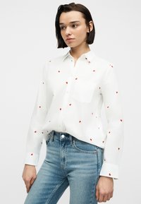 Νεαρή γυναίκα με κοντά σκούρα μαλλιά, φορώντας λευκό πουκάμισο με κόκκινα σχέδια καρδιών και μπλε τζιν, κοιτάζει προς τα πλάγια.