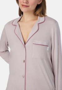 Vrouw in een lichtpaars pyjamashirt met contrasterende donkerpaarse randen, een borstzak en twee knopen, weergegeven van nek tot taille.