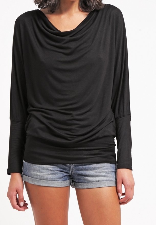 Femme portant un haut noir à manches longues drapé et un short en denim retroussé, debout devant un fond blanc.