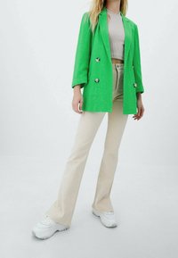 Femme portant un blazer vert vif, un haut court beige, un pantalon évasé beige et des baskets blanches, debout devant un fond uni.