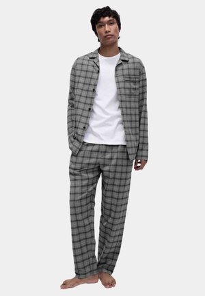 Junger Mann trägt grau kariertes Hemd und passende Hose über einem weißen T-Shirt, steht barfuß vor einem einfarbigen Hintergrund.