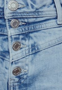 Jeans de mezclilla azul claro con botones de metal plateado grabados y detalles de costura visibles. La tela tiene una textura desgastada.