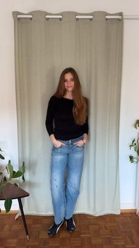 Czarna bluzka z długim rękawem, jasnoniebieskie dżinsy z przetarciami i kolorowymi plamami farby oraz czarne buty sportowe na tle beżowej zasłony.