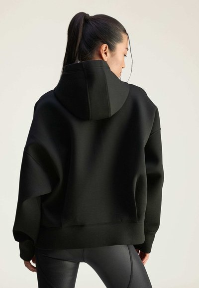 Čierna prešívaná mikina s vysokým golierom a kapucňou; mäkká, textúrovaná tkanina; voľný strih; zobrazená zozadu, kombinovaná s čiernymi legínami.