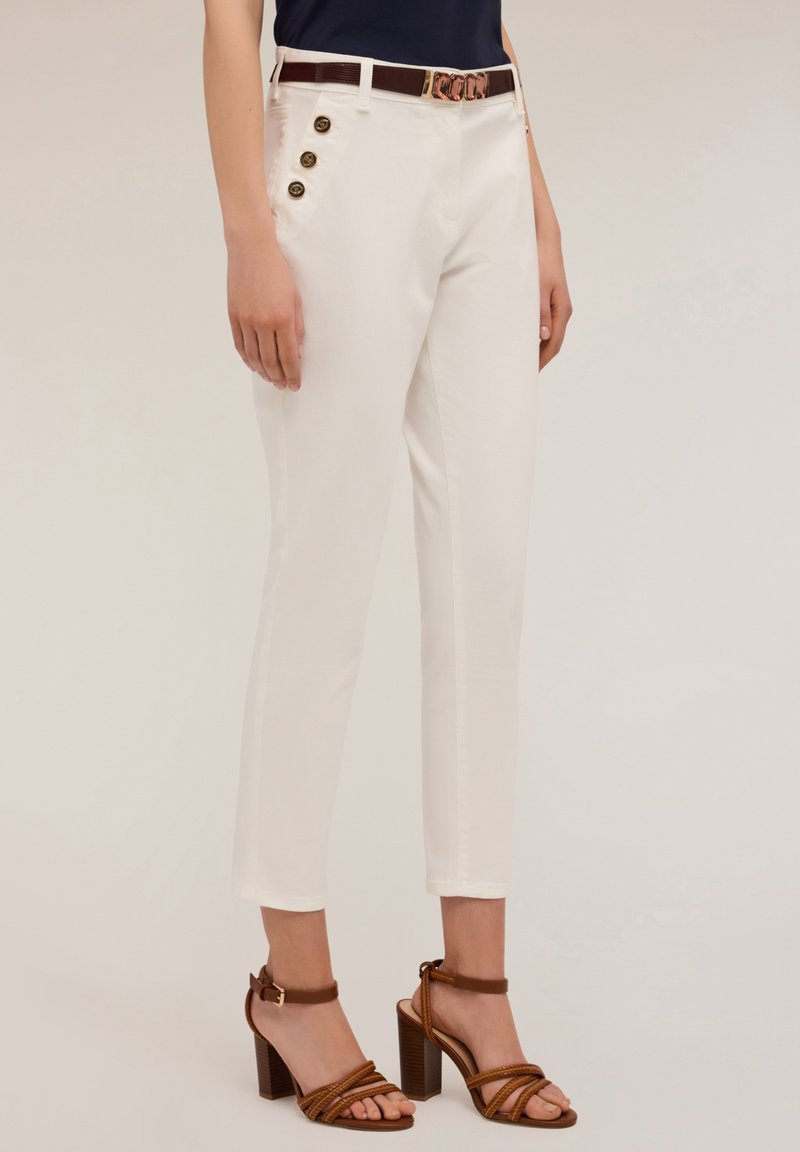 Pantalon blanc ajusté avec des boutons sur le côté, accompagné d'une ceinture marron. Le tissu semble lisse avec un léger éclat. Sandales à talons marron.