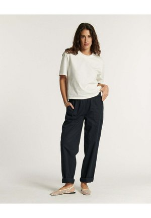 Femme debout, les mains dans les poches, portant une chemise blanche à manches courtes, un pantalon noir ample retroussé aux chevilles, et des chaussures plates tressées beige.