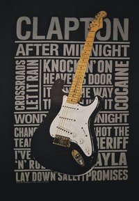 Zwarte T-shirt met een witte grafiek van een elektrische gitaar. Achtergrondtekst toont songtitels die geassocieerd worden met Eric Clapton in vetgedrukte letters.