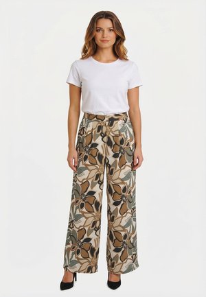 Donna in piedi che indossa una camicia bianca a maniche corte, pantaloni a gamba larga con motivo floreale in toni terrosi e tacchi alti neri.