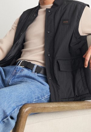 Homme assis sur une chaise rembourrée portant une veste noire sans manches, un pull beige, un jean bleu et une ceinture noire avec une boucle argentée.