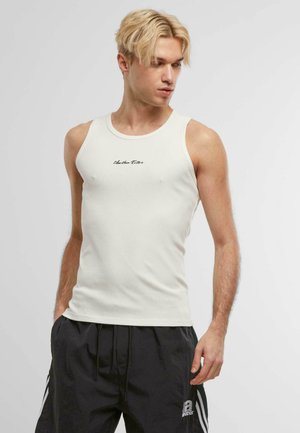 Hvid ribbet tanktop med en tætsiddende design og en rund halsudskæring, med sort tekst trykt på brystet. Båret med sorte shorts.
