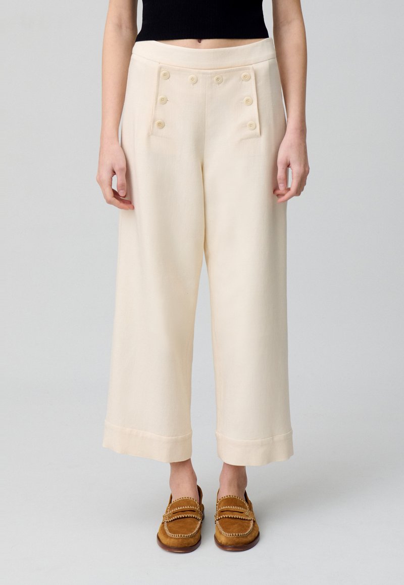 Pantalons larges crème avec des détails de boutons croisés, des poches avant et une texture lisse, associés à des mocassins en cuir marron.