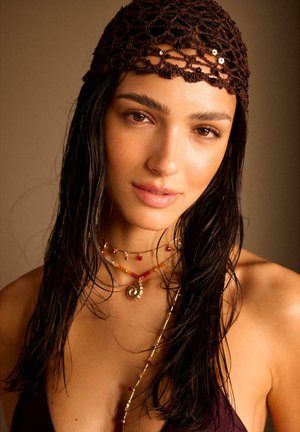 Kvinde med vådt mørkt hår iført en brun strikket pandebånd og lagdelte perlekæder med en varm neutral baggrund.