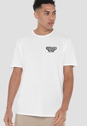 Man looking sideways wearing a white T-shirt with "Stranger Things Tales from 85" logo on the chest, standing against a plain background.