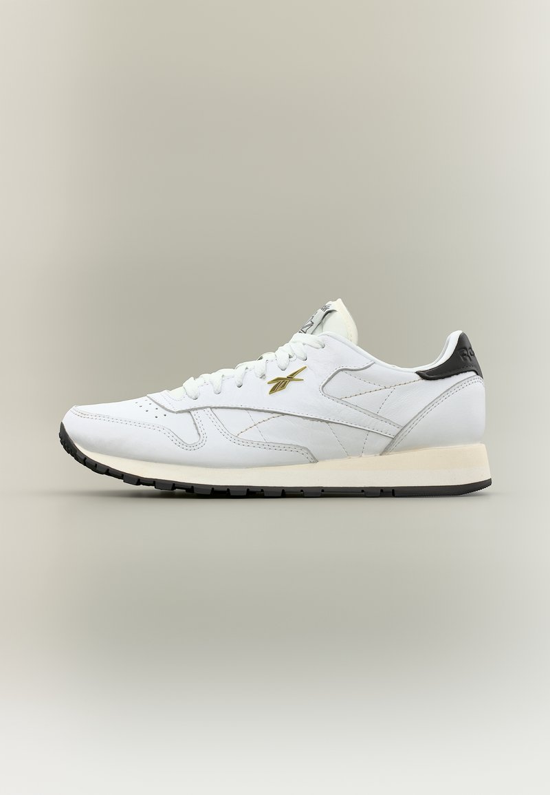 Baskets en cuir blanc avec semelle et languette de talon noires, logo doré sur le côté et lacets blancs, montrées de profil sur fond neutre.
