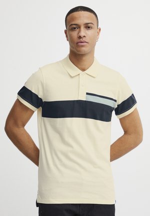 Polo skjorte i cremet bomuldsstof med en marineblå vandret stribe og en lyseblå accent på venstre side. Korte ærmer med ribkant.