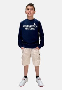 Felpa blu con la scritta "AERONAUTICA MILITARE", pantaloni cargo beige, sneakers bianchi con dettagli rossi, in piedi con le mani in tasca.