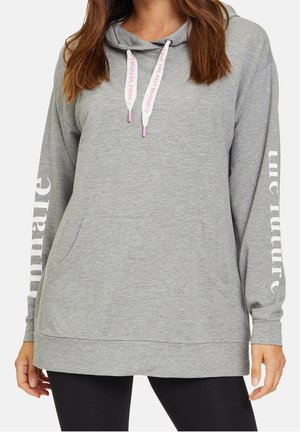 Kobieta nosząca jasnoszarą bluzę z kapturem z białym napisem „Inhale the future” na rękawach i różowym napisem na sznurków, w połączeniu z czarnymi legginsami.