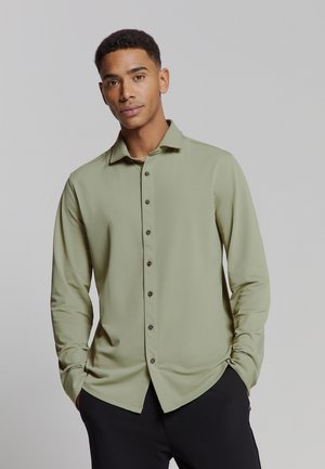 Homme avec des cheveux courts et bouclés, portant une chemise légère olivâtre à boutons et un pantalon noir, les mains dans les poches, se tenant devant un fond gris.
