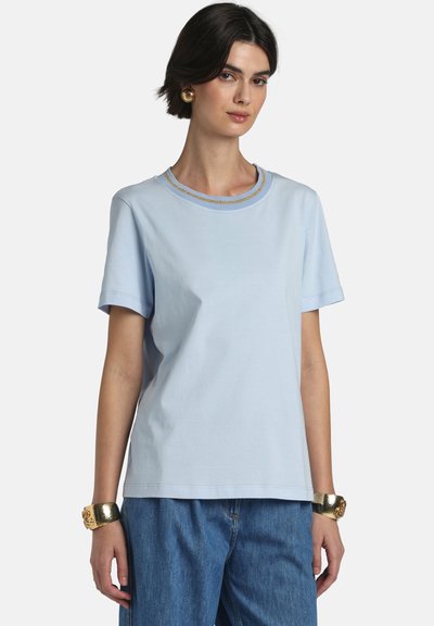 Kvinde iført en lyseblå T-shirt med korte ærmer og guldornamenteret halsudskæring, blå jeans, guldbøjler og guldbøjler på begge håndled.