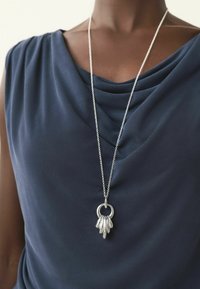 Zilveren ketting met een ronde hanger, voorzien van vijf langwerpige, taps toelopende charme, gedragen over een donkerblauwe gedrapeerde top.