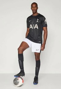 Mies, oblečený v černém fotbalovém dresu Tottenham Hotspur, stojí s jednou nohou na černo-bílém fotbalovém míči na jednobarevném pozadí.