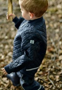 Chaqueta acolchada azul marino con cuello acanalado y detalles en blanco, usada por un niño que sostiene un palo, en un entorno de suelo forestal.